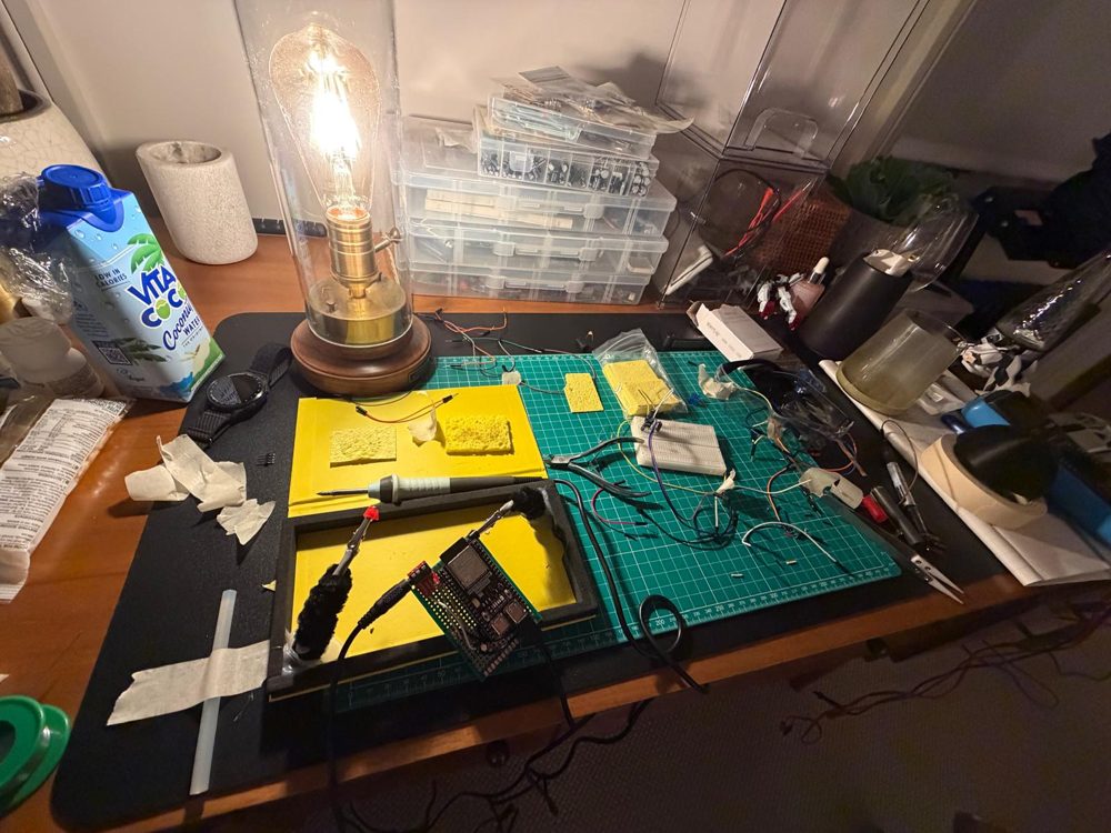 My soldering bench: iron, cutting mat, component boxes and homemade helping hands holding the board.