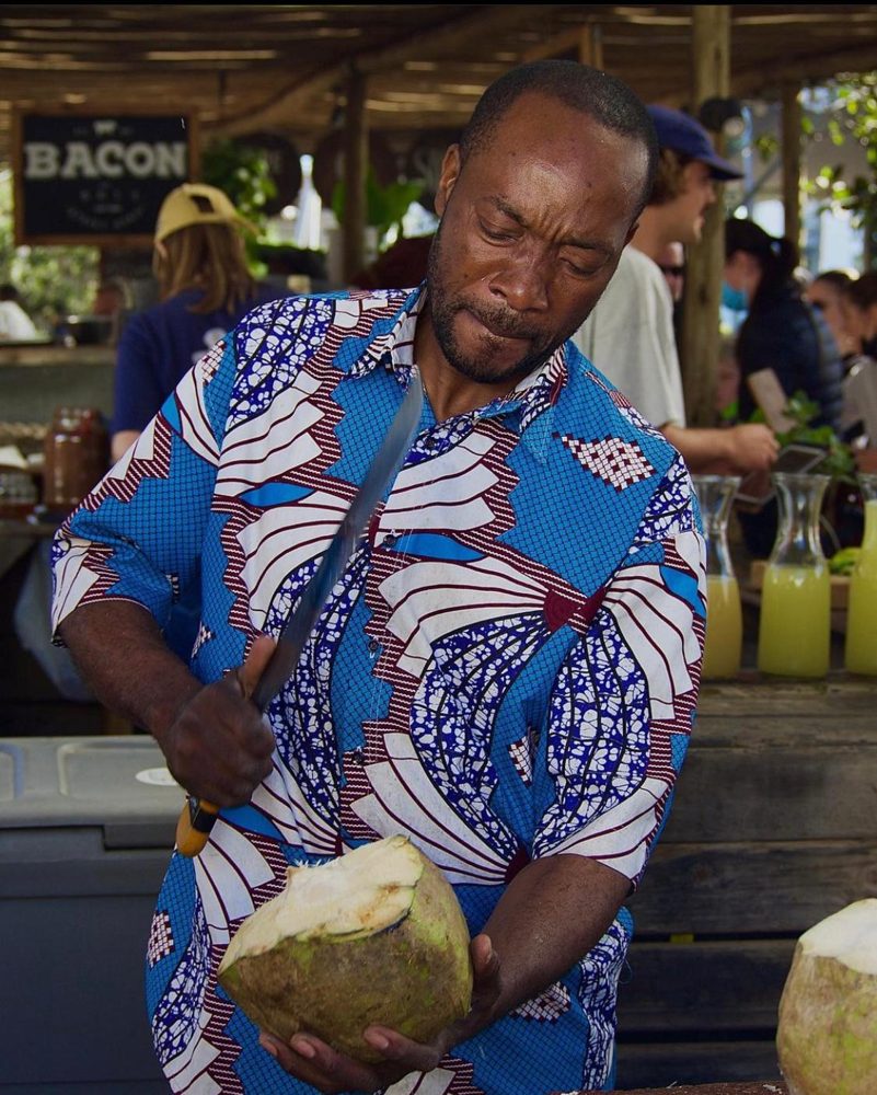 A man cutting open a coconut with a machete at a market stall.